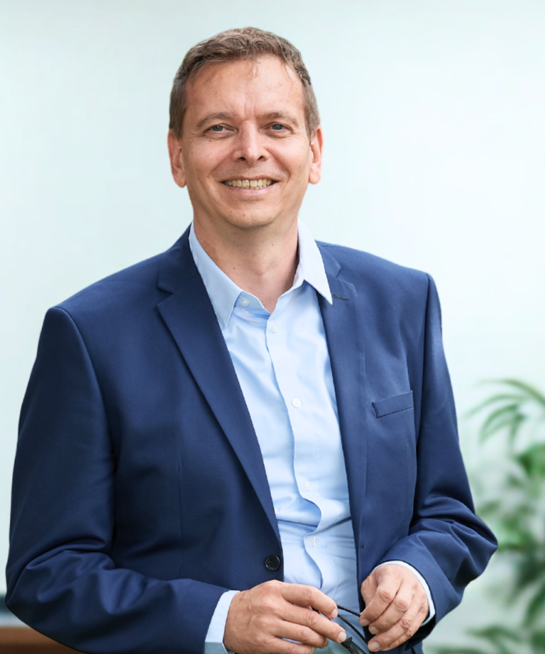 Dr. Frank Jürgensen in Anzug lächelt, steht in einem hellen Raum mit Pflanzen im Hintergrund. Dr. Frank Jürgensen in Anzug lächelt, steht in einem hellen Raum mit Pflanzen im Hintergrund.