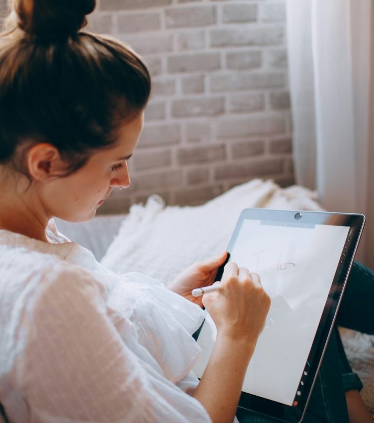 Frau, die auf einem Tablet schreibt, sitzt auf einem Sofa mit gemusterter Rückwand. Frau, die auf einem Tablet schreibt, sitzt auf einem Sofa mit gemusterter Rückwand.