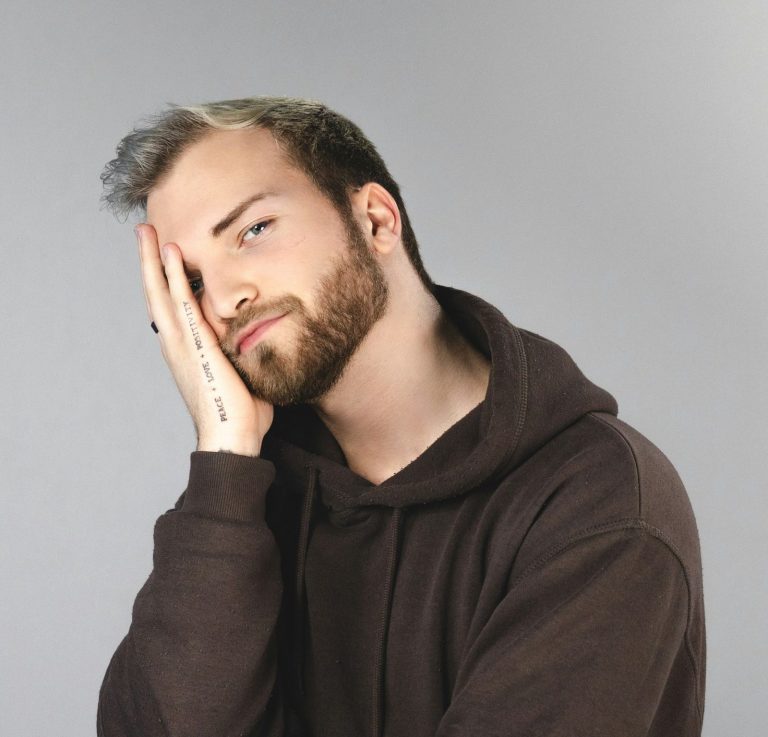 Junger hochsensibler Mann mit Bart, der nachdenklich in die Kamera schaut und seinen Kopf mit einer Hand stützt. Junger Mann mit Bart, der nachdenklich in die Kamera schaut und seinen Kopf mit einer Hand stützt.